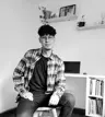 Roiber, sentado frente a su escritorio en un entorno de trabajo minimalista, mirando a la cámara con una expresión serena. Lleva gafas, una camiseta negra y una camisa de cuadros. Al fondo se observan libros, una laptop y estantes con plantas y decoraciones.