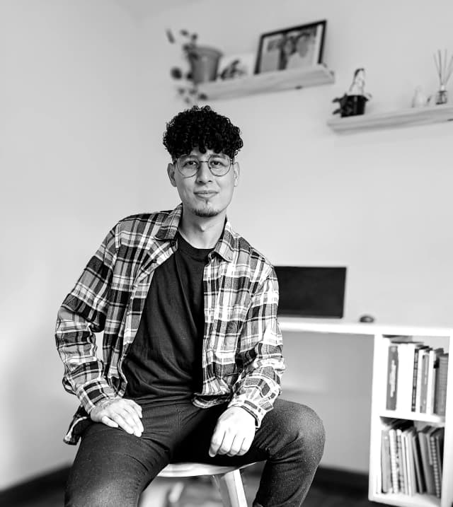 Roiber, sentado frente a su escritorio en un entorno de trabajo minimalista, mirando a la cámara con una expresión serena. Lleva gafas, una camiseta negra y una camisa de cuadros. Al fondo se observan libros, una laptop y estantes con plantas y decoraciones.